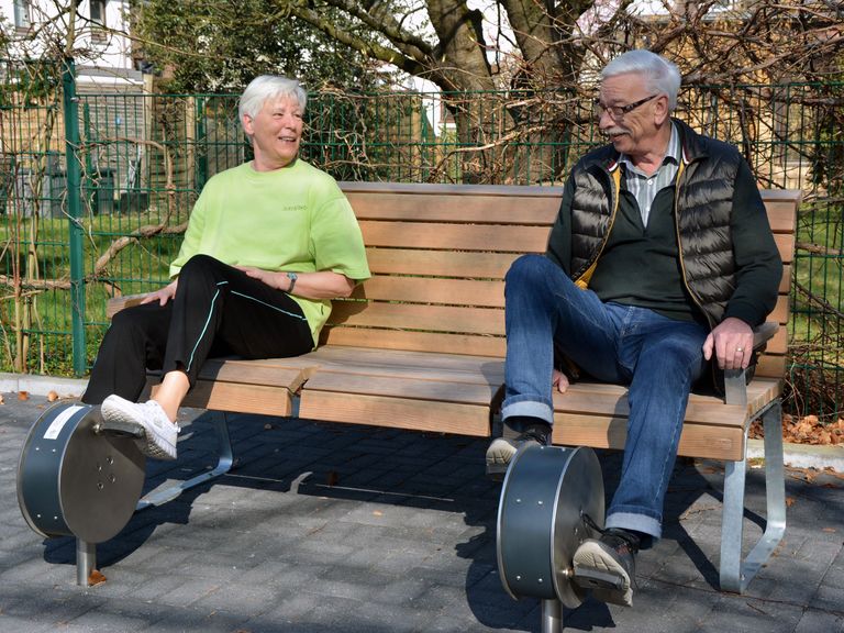 Ein älteres Paar trainiert auf einer Bank sitzend mit speziellen Geräten die Beine.