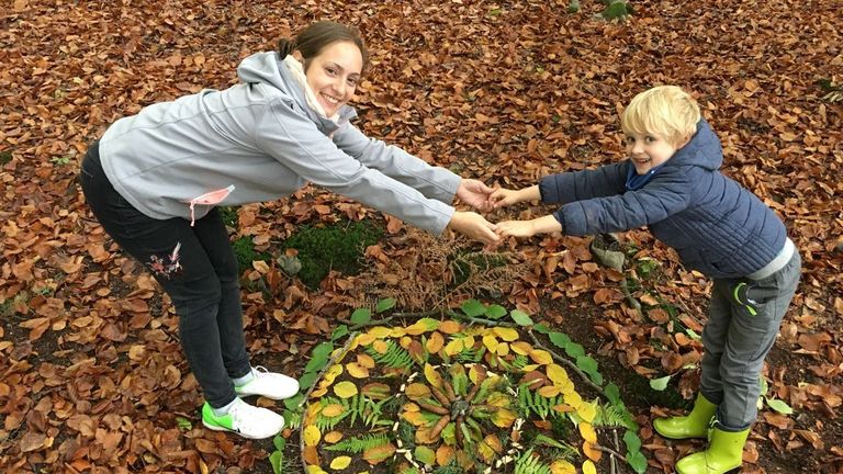 Mutter und Sohn halten sich an den Händen über einem Kunstwerk aus Laub und Zweigen.